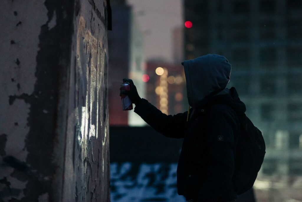 A hooded figure paints graffiti on a wall.